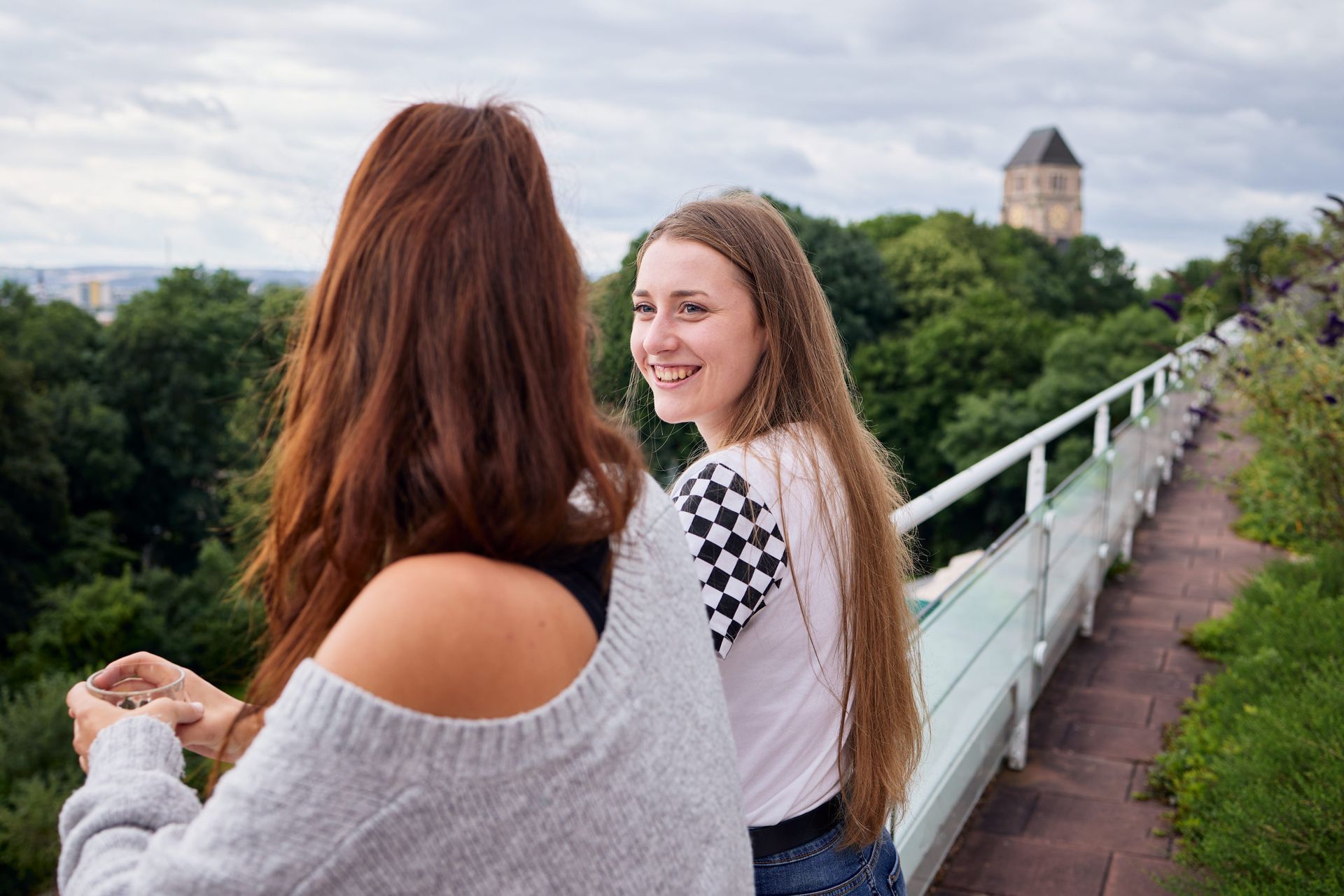 Zwei junge Frauen auf einem Steg mit Blick auf eine üppig grüne Landschaft. Eine lächelt zurück. Ein Steinturm ist zu sehen.