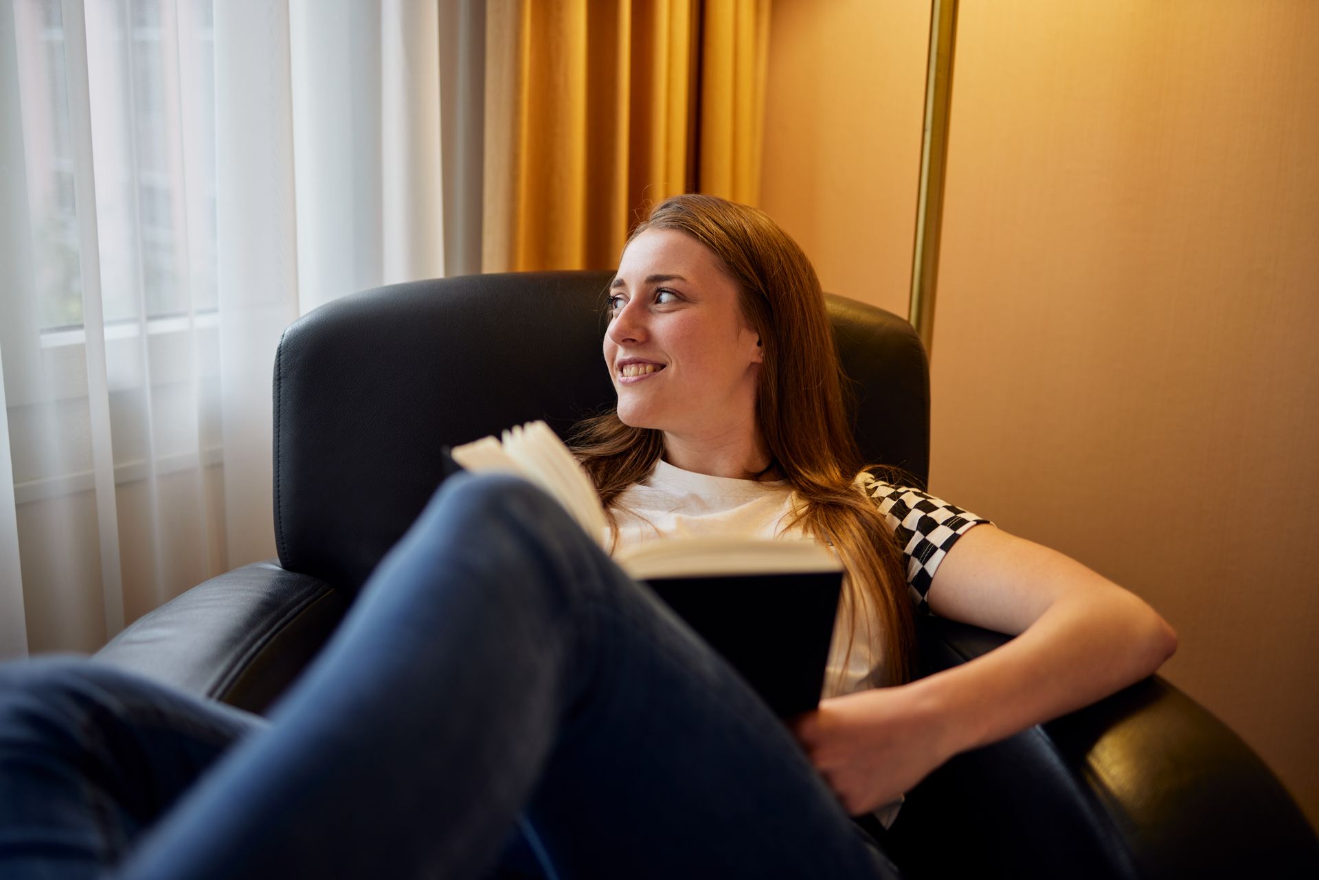 Eine Frau im Sessel schaut mit einem Buch in der Hand aus dem Fenster. Sie trägt ein weißes Hemd und Jeans.