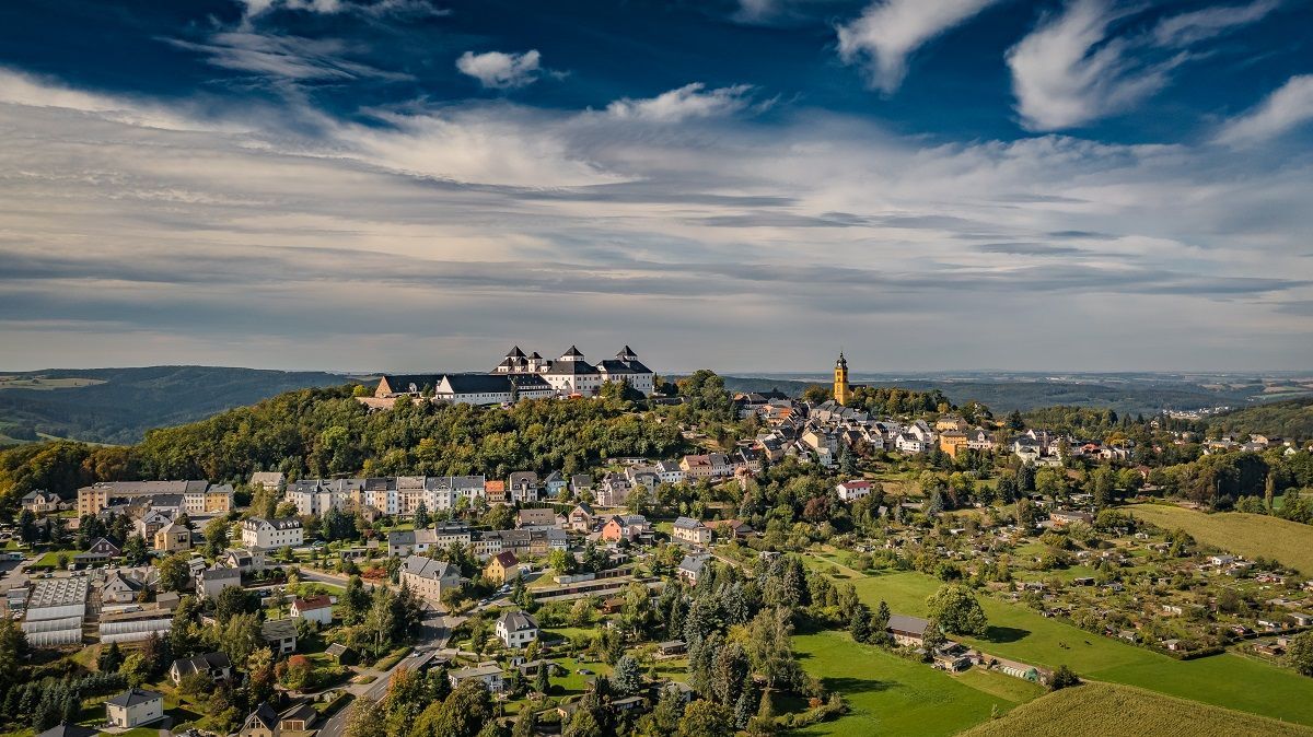 Stadtbild mit einer Burg auf einem Hügel, umgeben von Häusern, Bäumen und grünen Feldern unter einem bewölkten blauen Himmel.