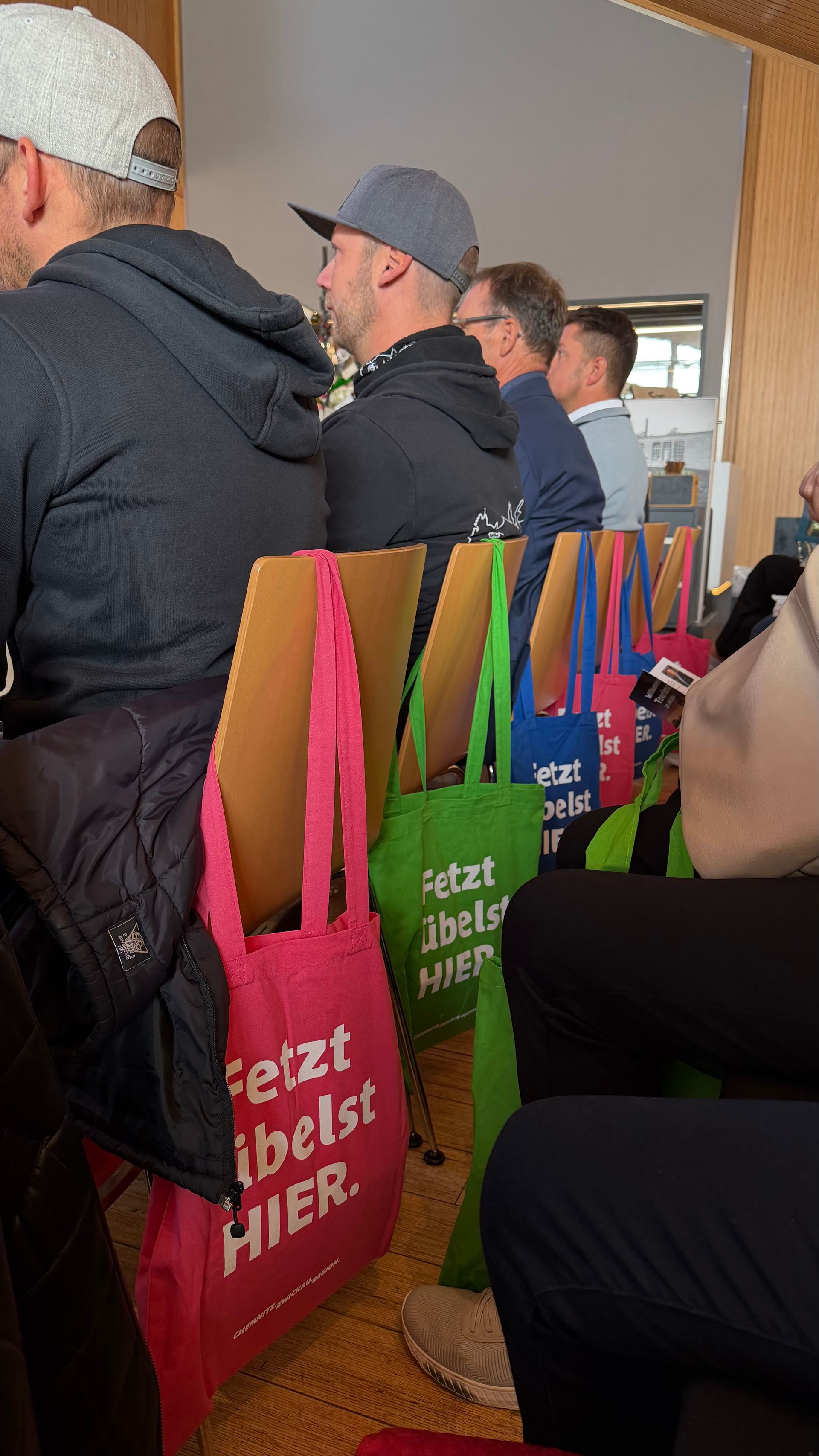 Menschen, die beim 2. Tourismustag sitzen, auf ihren Stühlen hängen bunte Beutel
