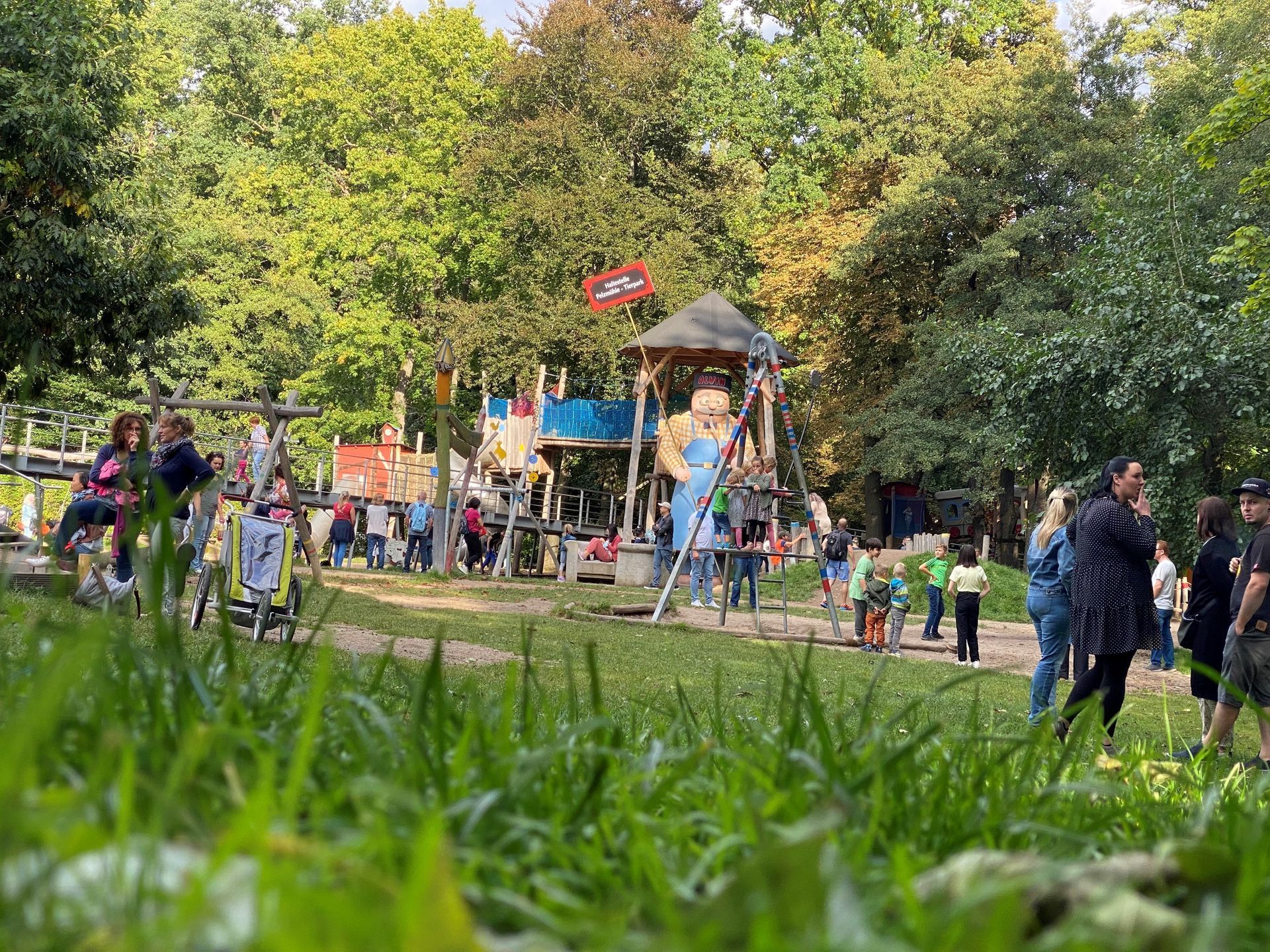 Menschen in einem Park in der Nähe eines Spielplatzes, im Vordergrund sind Bäume und Gras zu sehen.