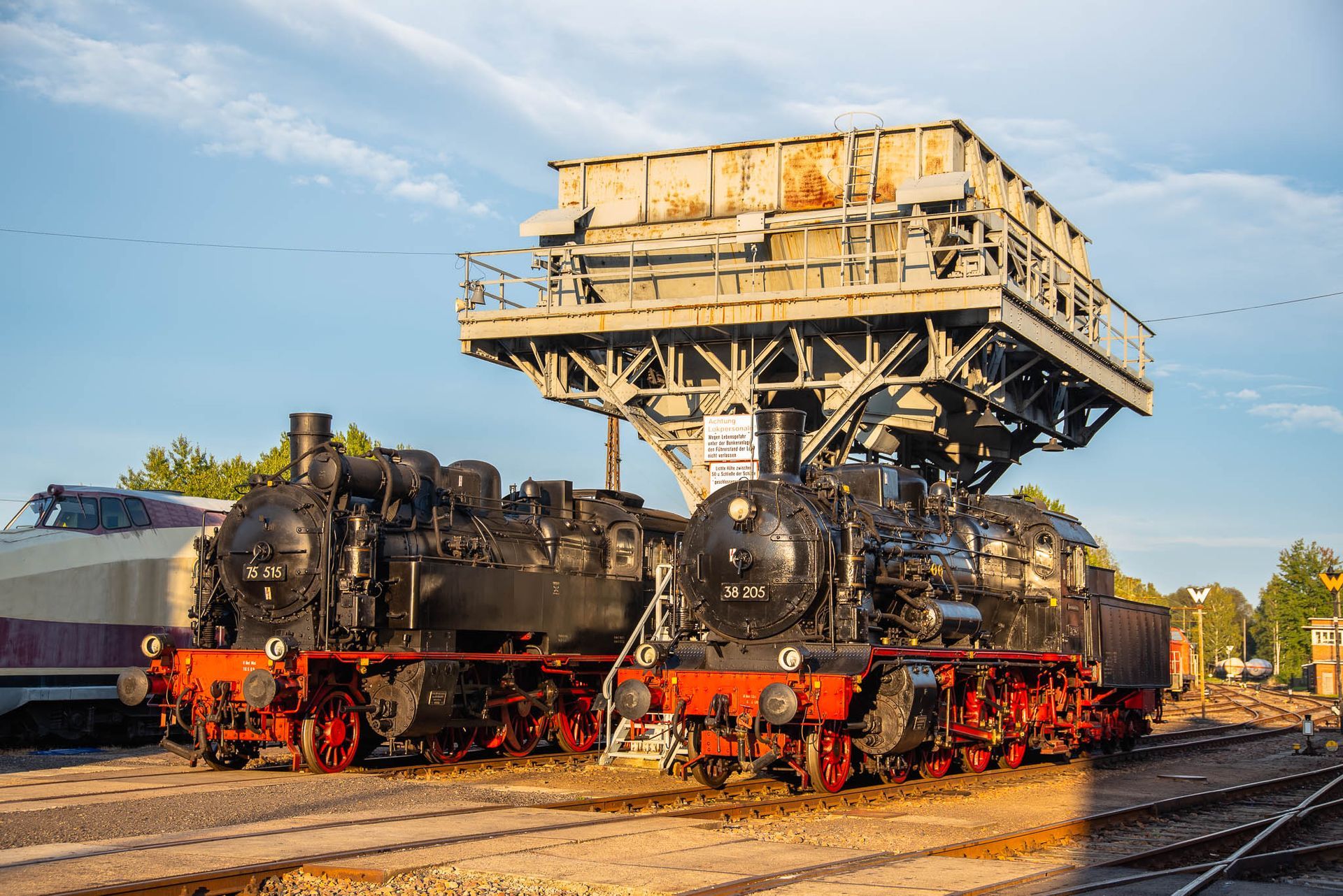 Schwarze Dampflokomotive unter einer großen industriellen Verladeanlage auf einem Rangierbahnhof.