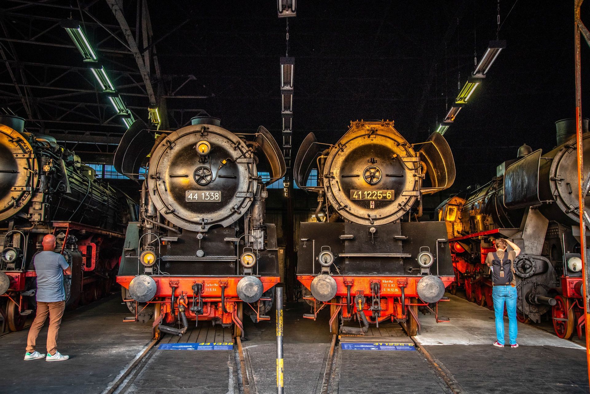 Drei historische Dampflokomotiven sind in einer dunklen Bahnhalle ausgestellt, daneben stehen Menschen