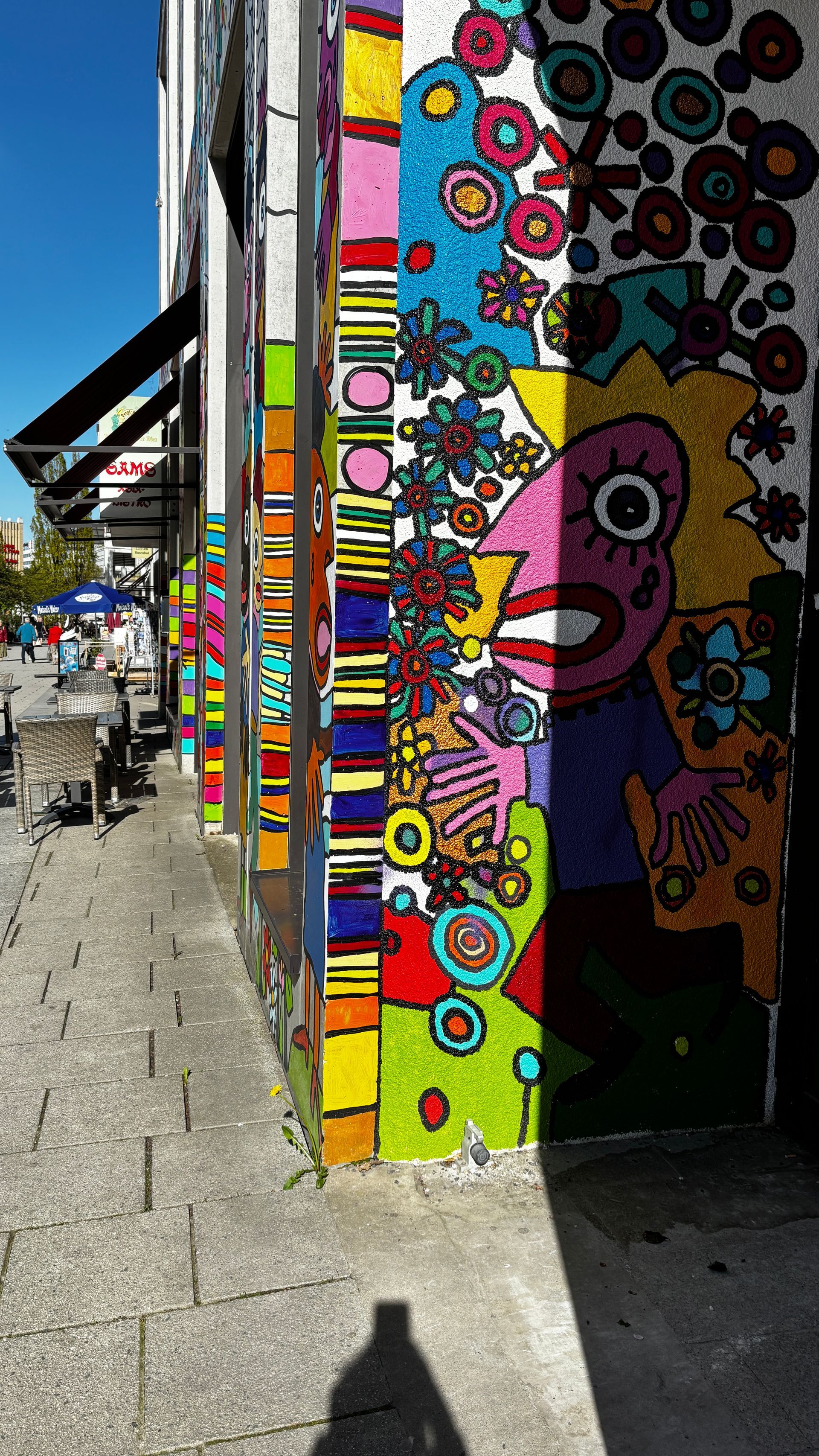 Farbenfrohes Graffiti-Wandbild auf einem städtischen Bürgersteig neben einem Gebäude, mit leuchtenden abstrakten Mustern und einem Comic-Gesicht.