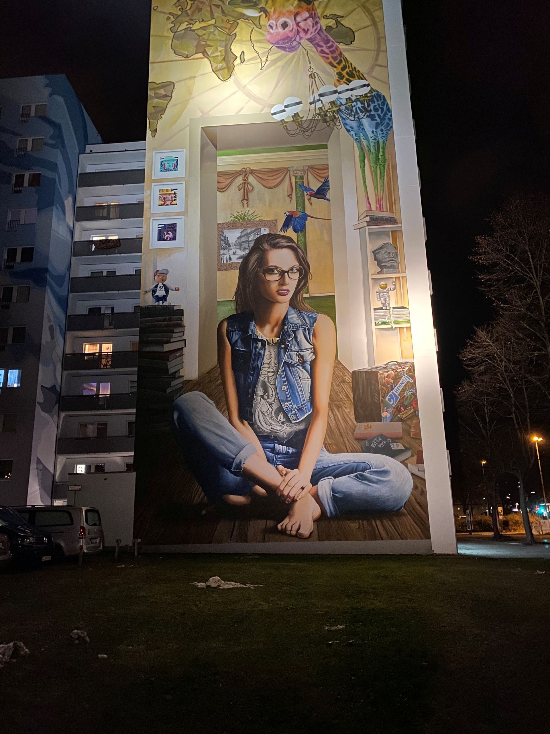 Großes Wandgemälde einer sitzenden Frau an einer Gebäudewand bei Nacht, beleuchtet von Straßenlaternen