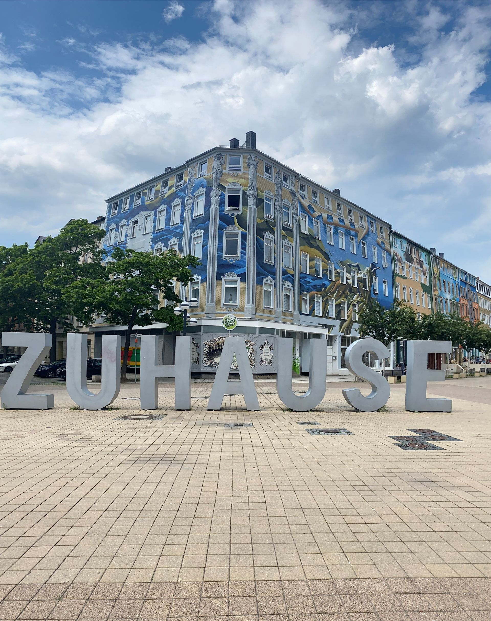 Große „ZUHAUSE“-Buchstaben vor einem mit einem blauen Wandgemälde verzierten Gebäude unter einem bew