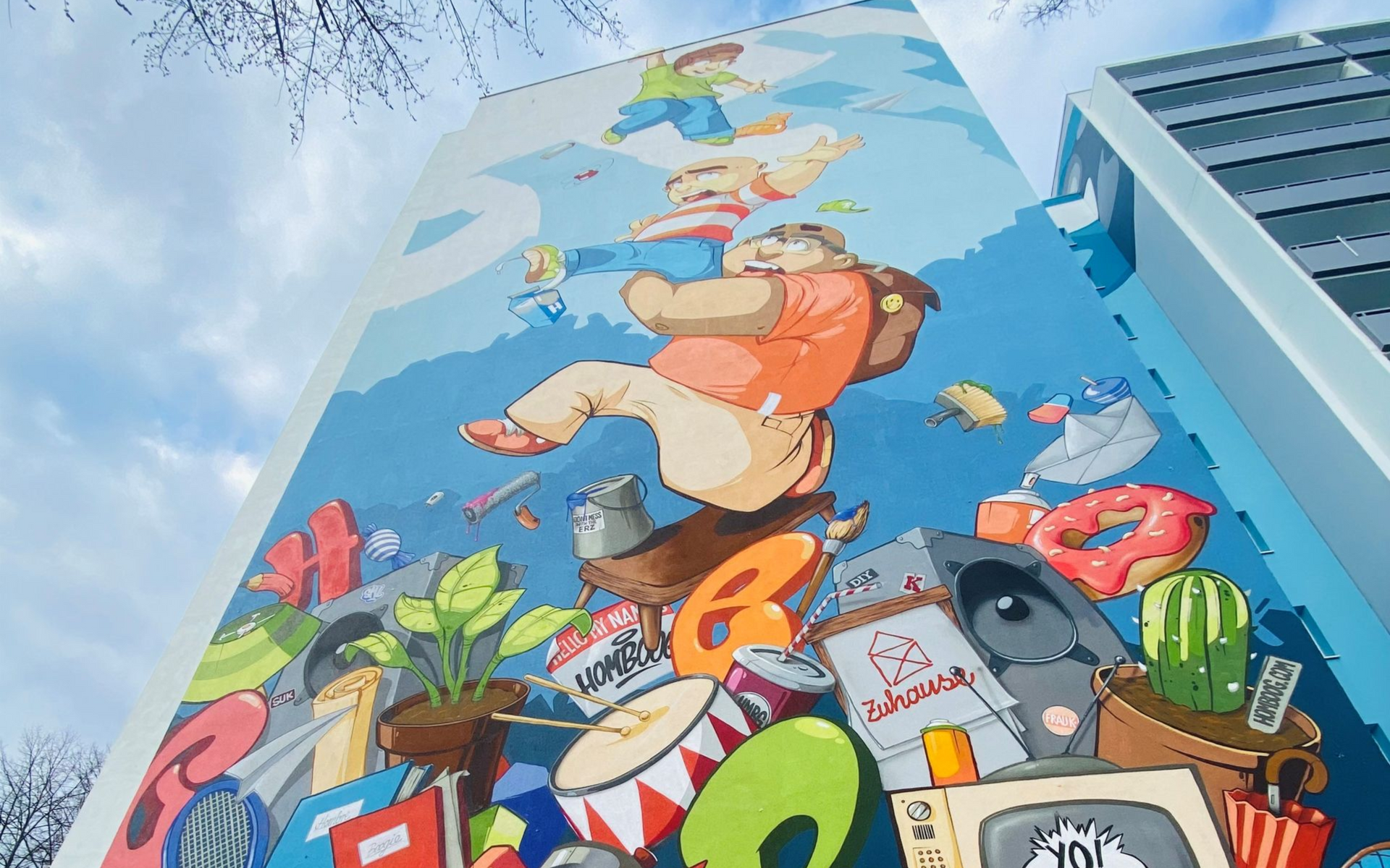 Farbenfrohes Wandgemälde an der Fassade eines hohen Gebäudes vor blauem Himmel, mit Comicfiguren und skurrilen Objekten