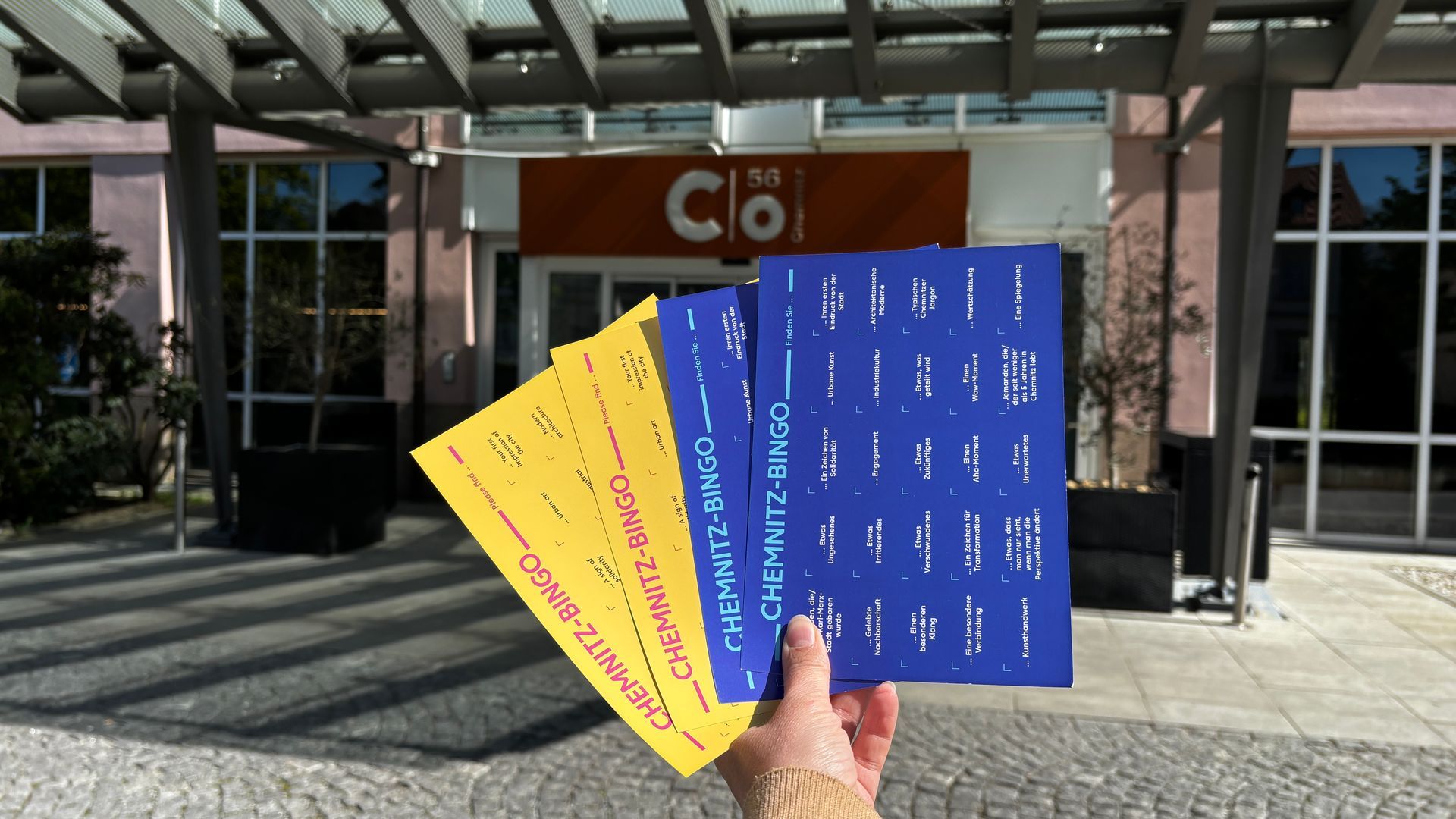 Eine Hand, die bunte Broschüren vor dem Eingang eines Gebäudes mit Glastüren und Stufen hält.