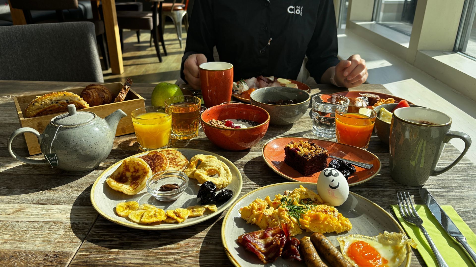 Ein Frühstücksbuffet mit Tellern voller Eier, Speck, Obst, Kaffee und Saft steht neben zwei sitzenden Personen.