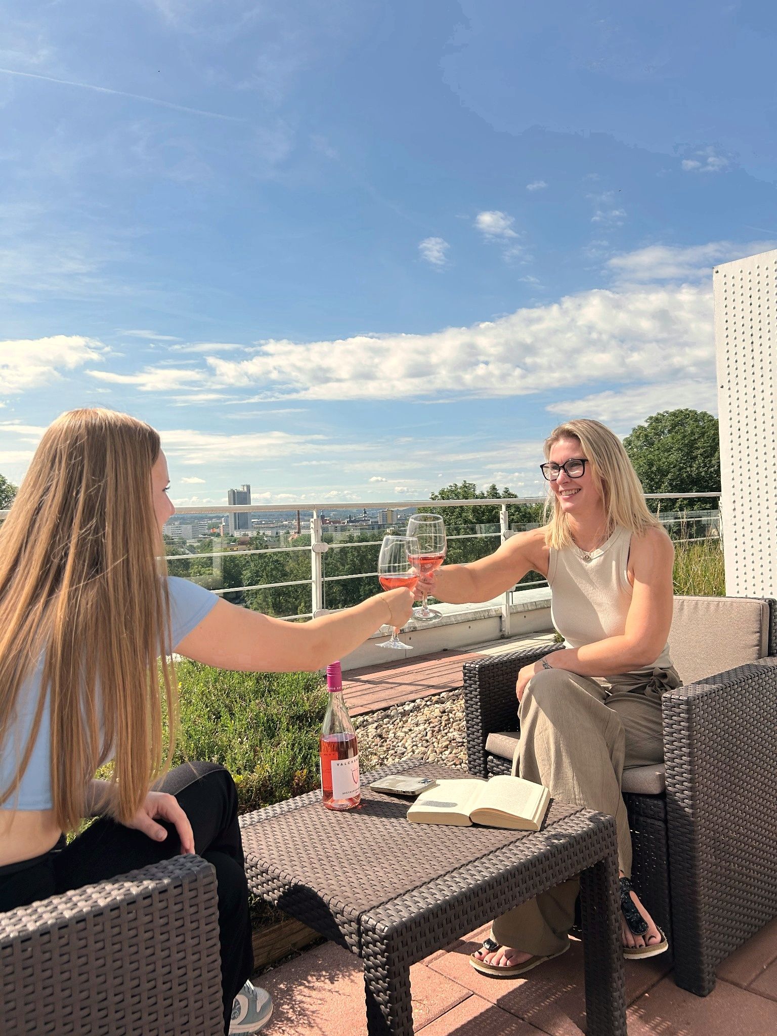 Zwei Personen stoßen auf einer Dachterrasse an, im Hintergrund die Skyline der Stadt und blauer Himmel.
