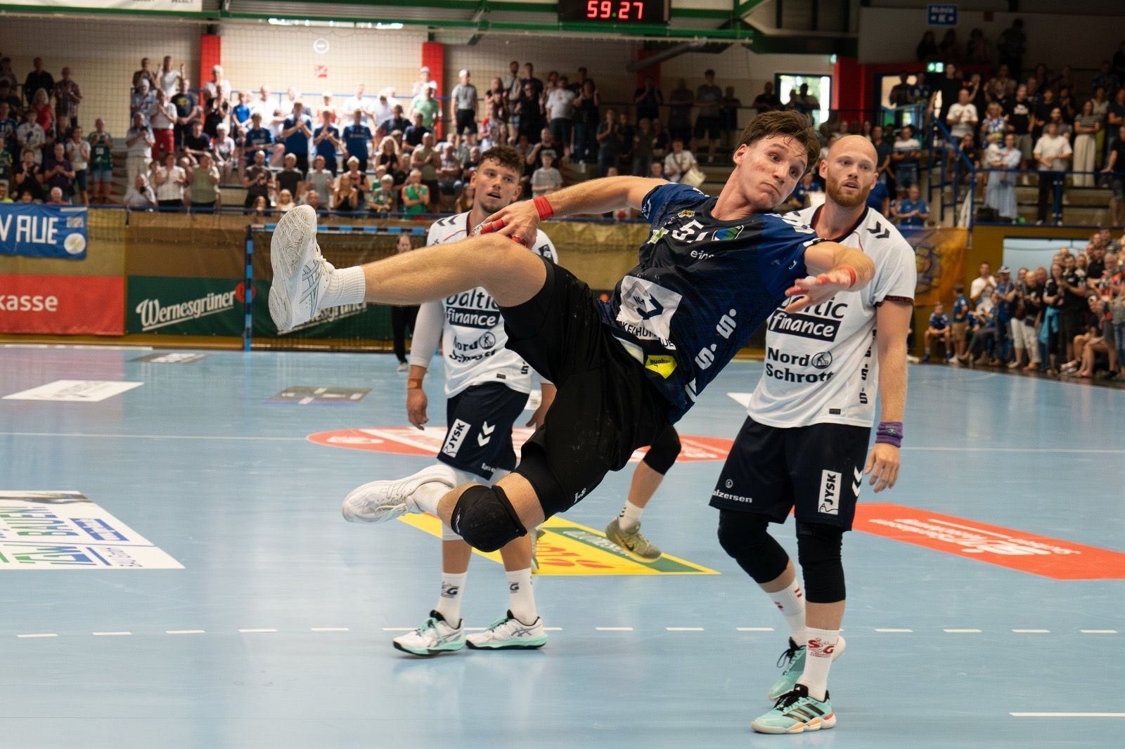 Handballspieler in schwarzer Kleidung, der den Ball in der Luft wirft, während er von zwei Spielern auf einer Hallenbahn verteidigt wird