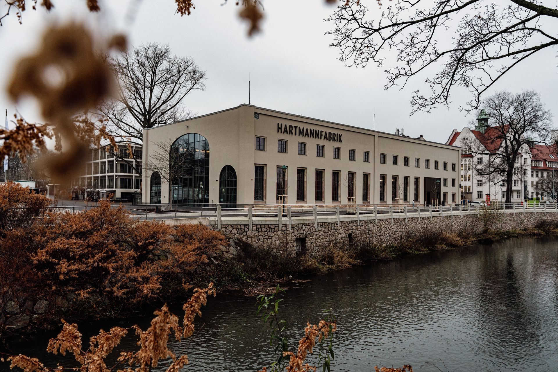 Die Hartmannfabrik, umgeben von kahlen Bäumen und Herbststräuchern unter grauem Himmel