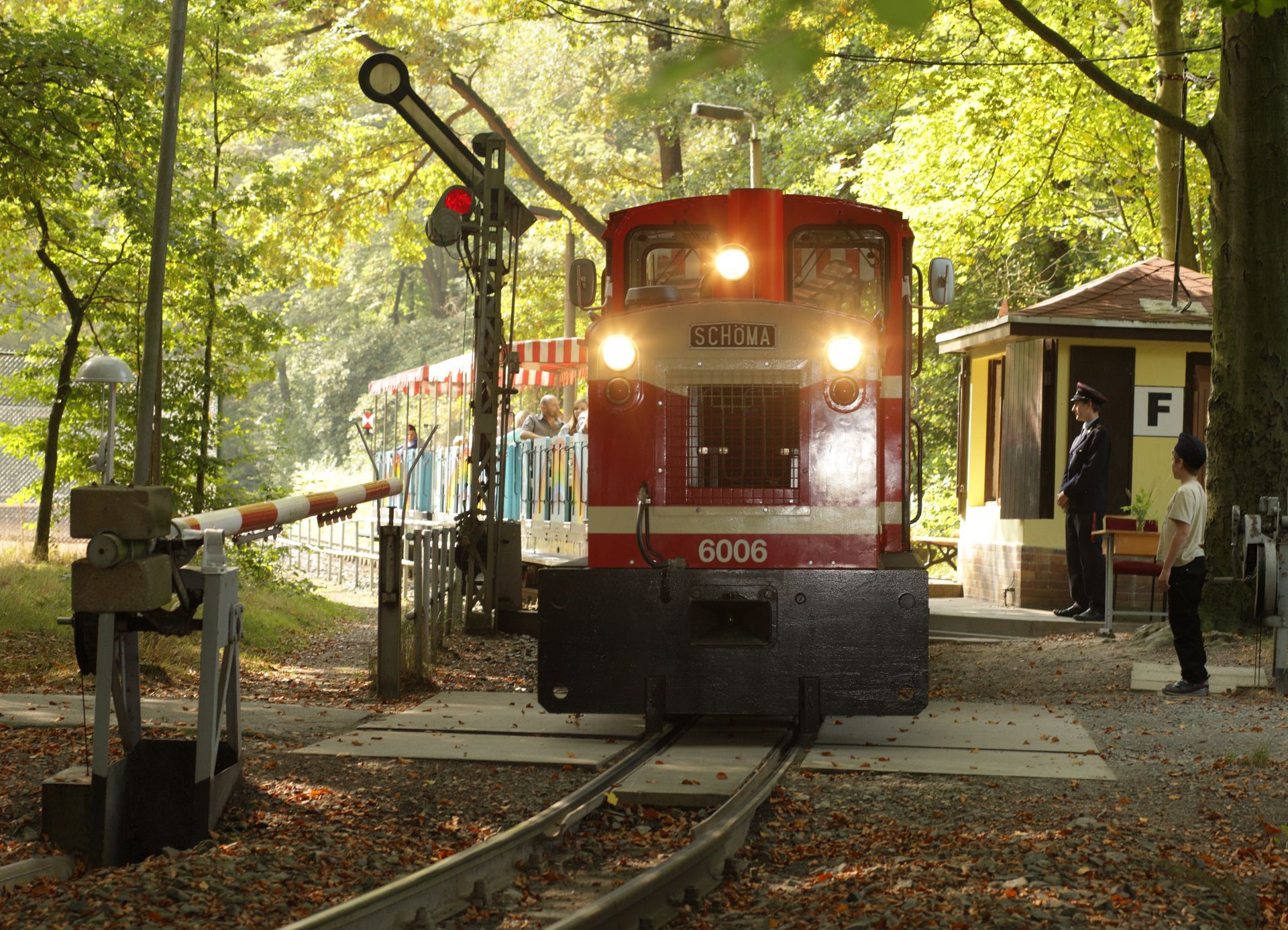 Ein roter Zug an einem bewaldeten Bahnübergang mit geschlossenen Schranken und eingeschalteten Scheinwerfern, umgeben von Bäumen und Menschen in der Nähe