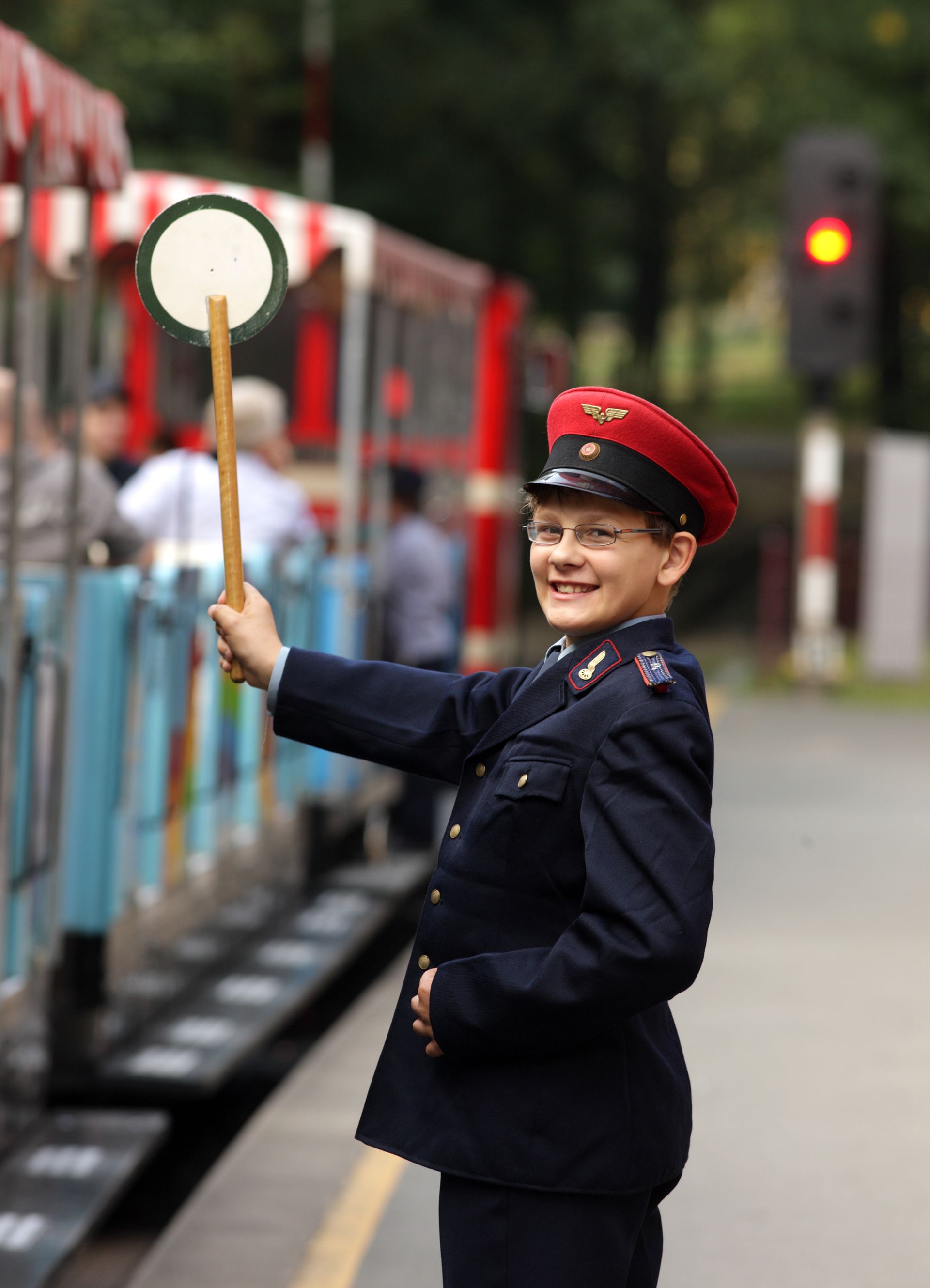 Ein Bahnmitarbeiter mit roter Mütze, der neben einem Bahnsteig Signale gibt, ein rundes Signalbrett