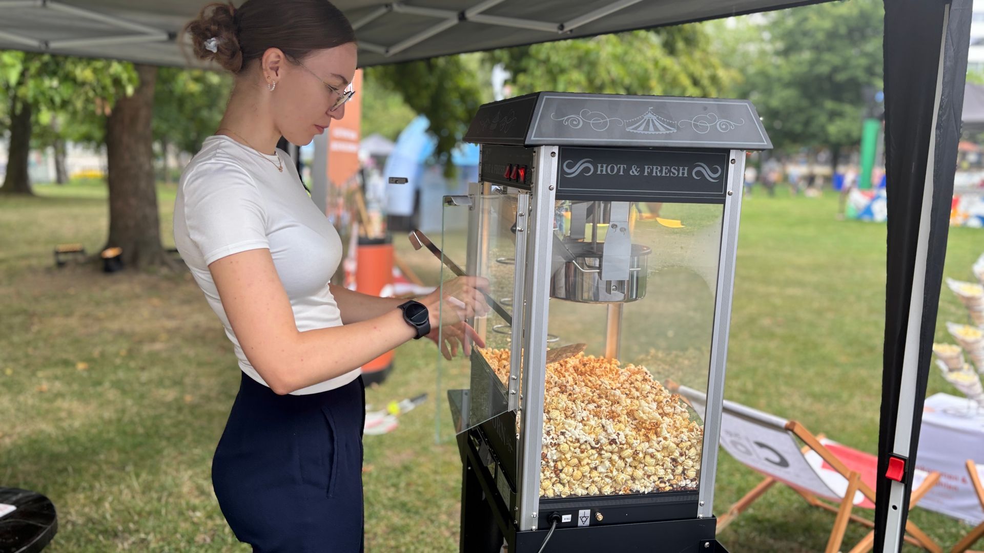 Eine Frau an einer Popcornmaschine im Freien unter einem Vordach, die bei einer Veranstaltung im Park Popcorn austeilt.