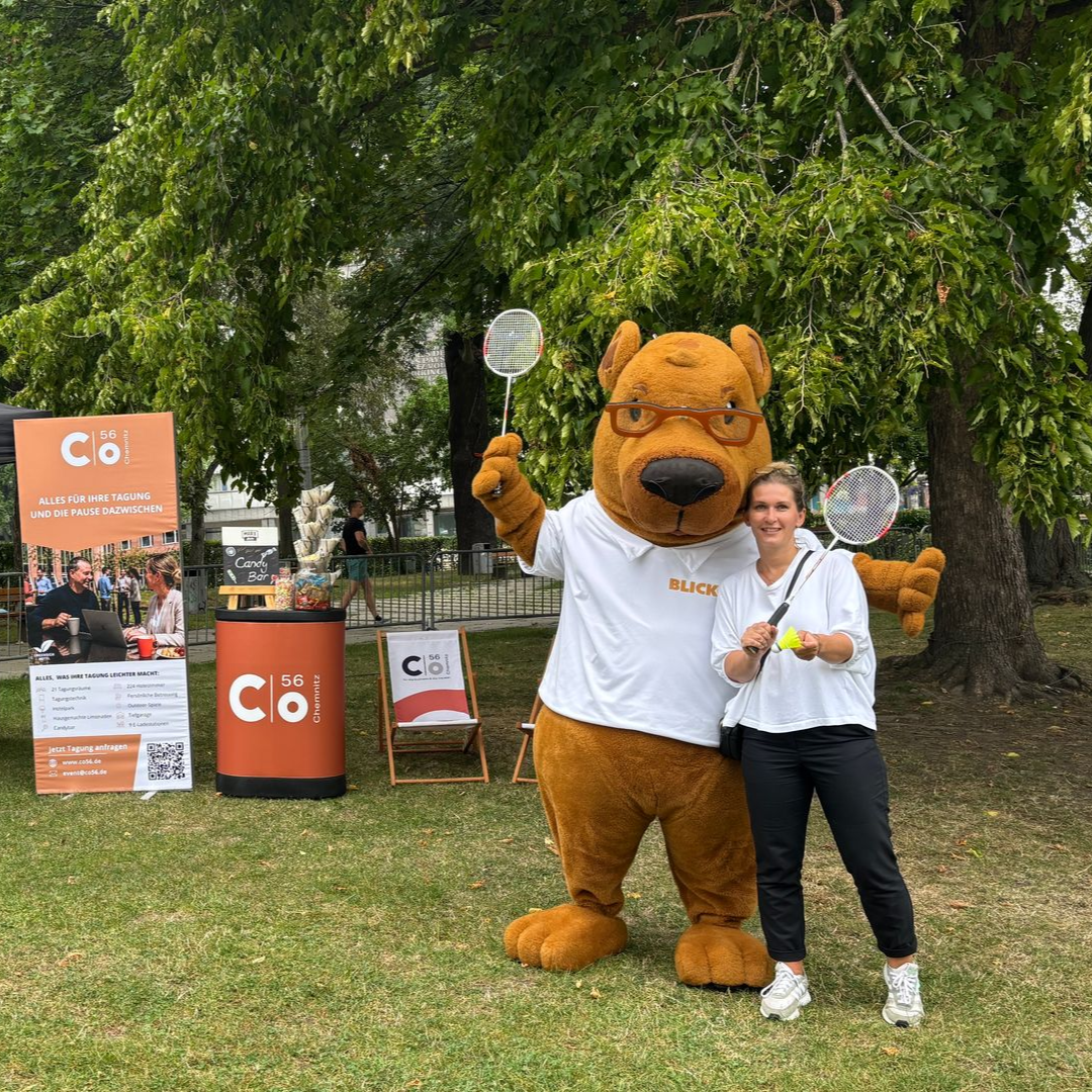 Ein Maskottchen in einem braunen Kostüm posiert mit einer Person an einem Stand im Freien unter einem Baum.