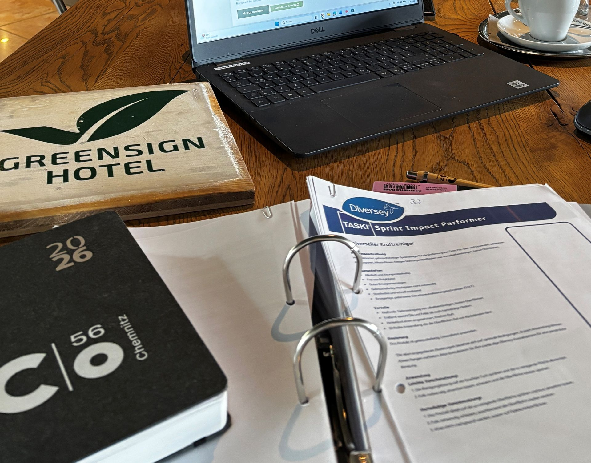 Ein Laptop auf einem Cafétisch mit Speisekarten, einer Kaffeetasse und einer modernen Hotellobby im Hintergrund.