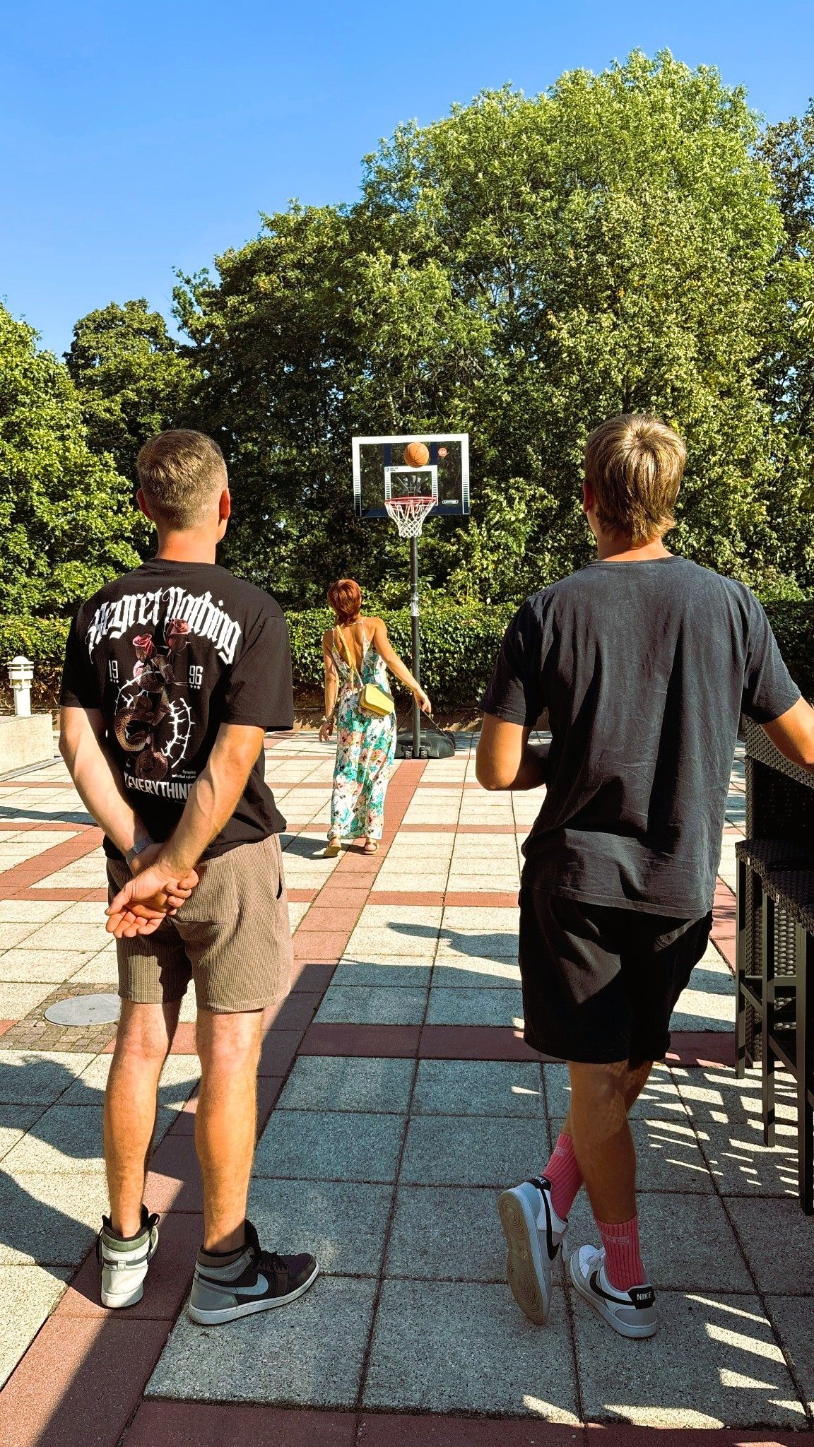 Zwei Personen gehen auf einen Basketballkorb auf einem sonnigen Freiplatz zu, im Hintergrund sind Bäume zu sehen.