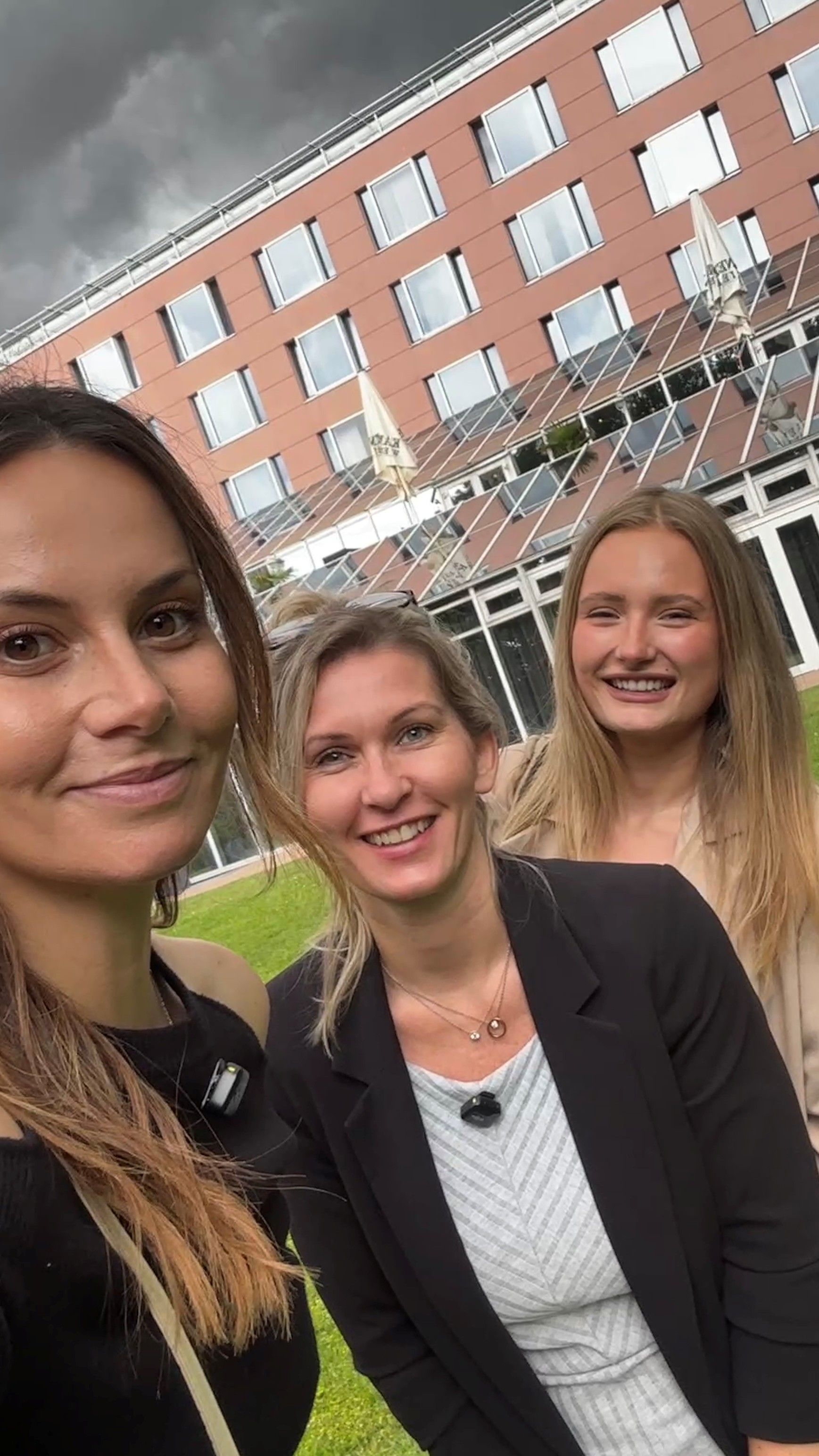 Drei Frauen, die im Freien vor einem Backsteingebäude und einer Rasenfläche mit weißen Gitterkonstruktionen lächeln.