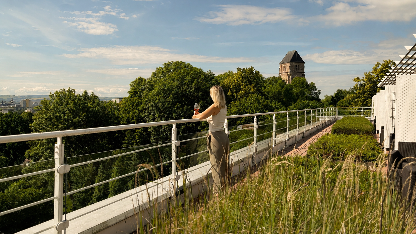 Eine Person steht auf einer Dachterrasse und blickt bei Sonnenuntergang auf eine grüne Landschaft und einen entfernten Turm.