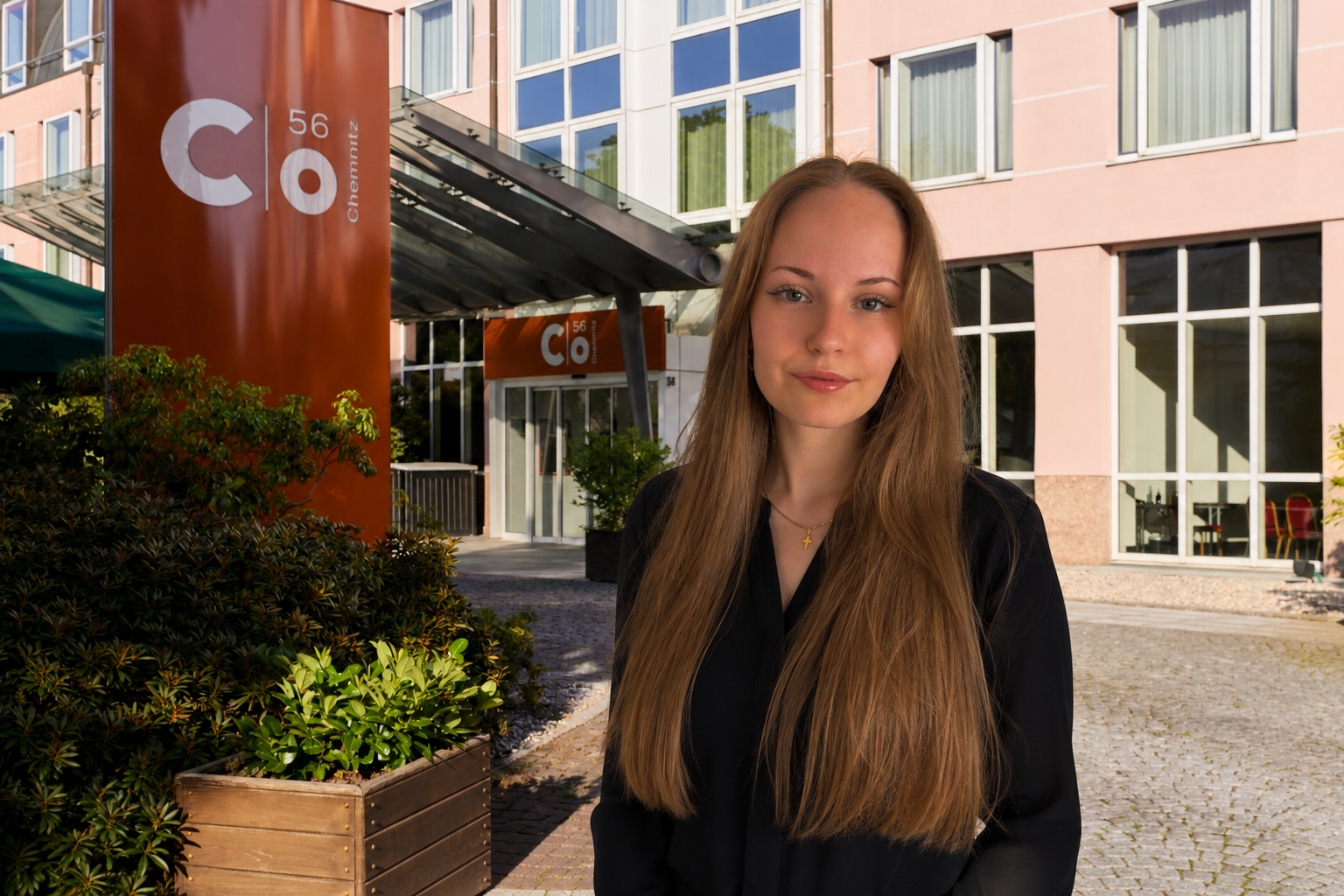 Eine Frau steht vor dem Eingang eines modernen Gebäudes, an dem ein rotes Schild mit der Aufschrift „C/o56 Chemnitz“ angebracht ist.