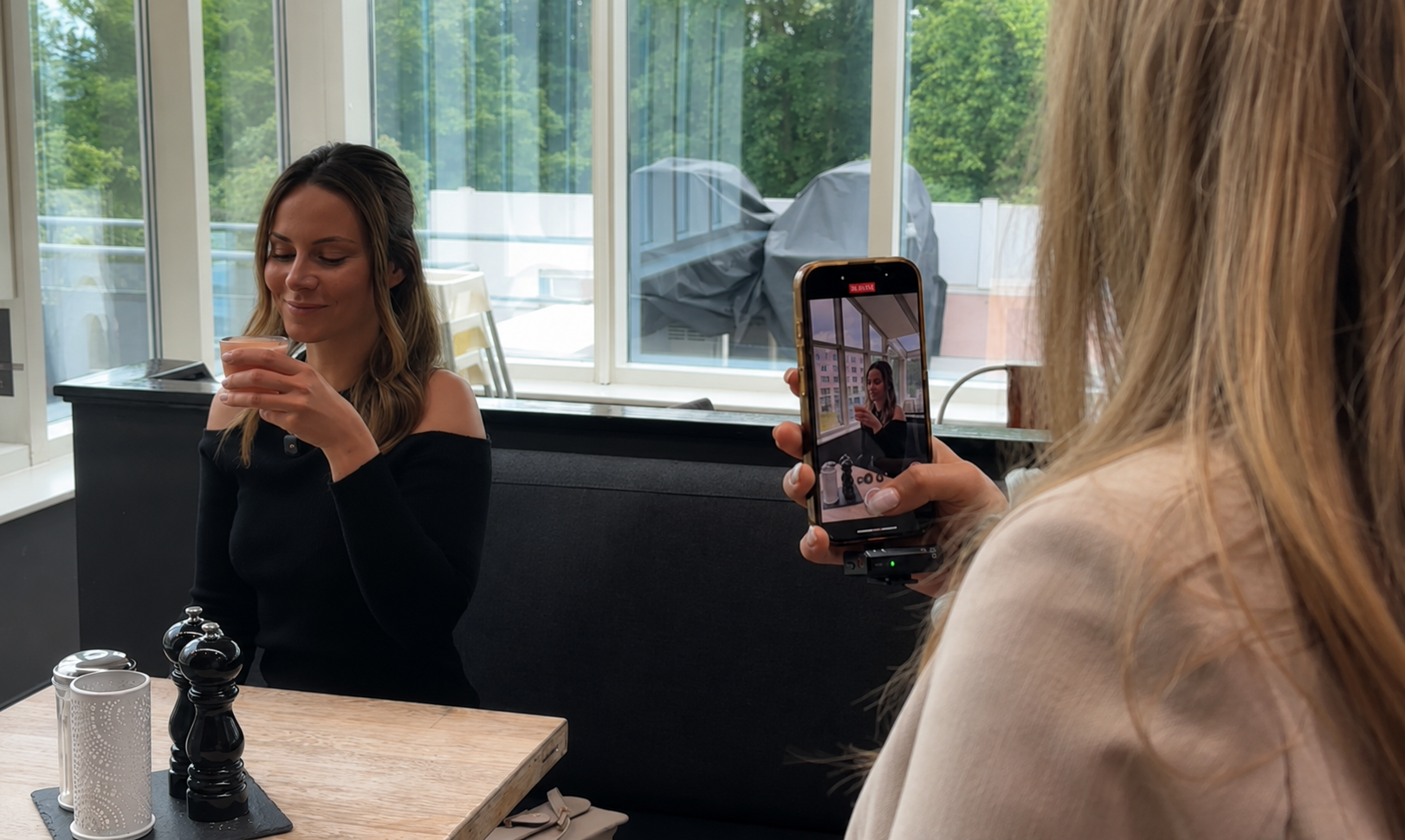 Eine Frau lächelt auf einer Cafébank, während eine andere Person sie mit dem Handy filmt.