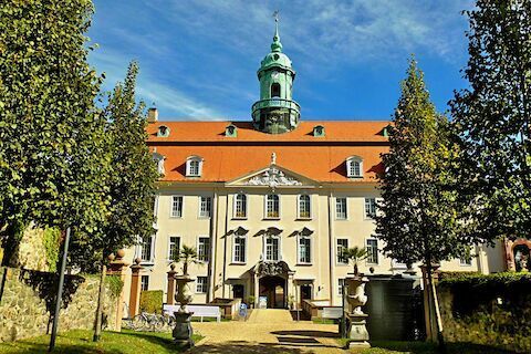 Beiges Gebäude mit rotem Dach, grünem Turm und davorliegenden Bäumen vor blauem Himmel.