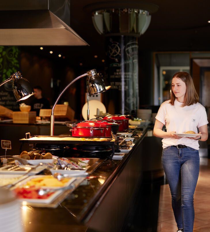 Frau steht am Buffet und hält einen Teller. In einem Restaurant stehen Tabletts mit Speisen unter Wärmelampen an der Theke.