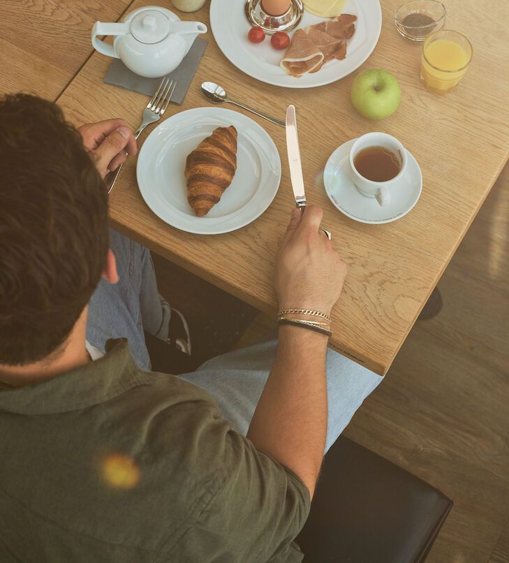 Person an einem Tisch mit Frühstück: Croissant, Eier, Speck, Obst, Tee.