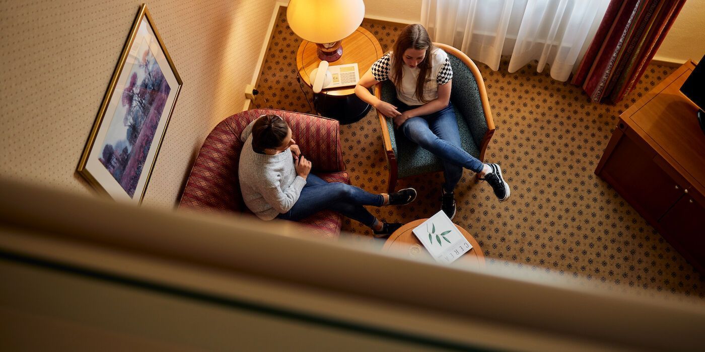 Zwei Personen sitzen in einem Hotelzimmer. Sie unterhalten sich; vor ihnen steht ein kleiner Tisch mit Papieren.