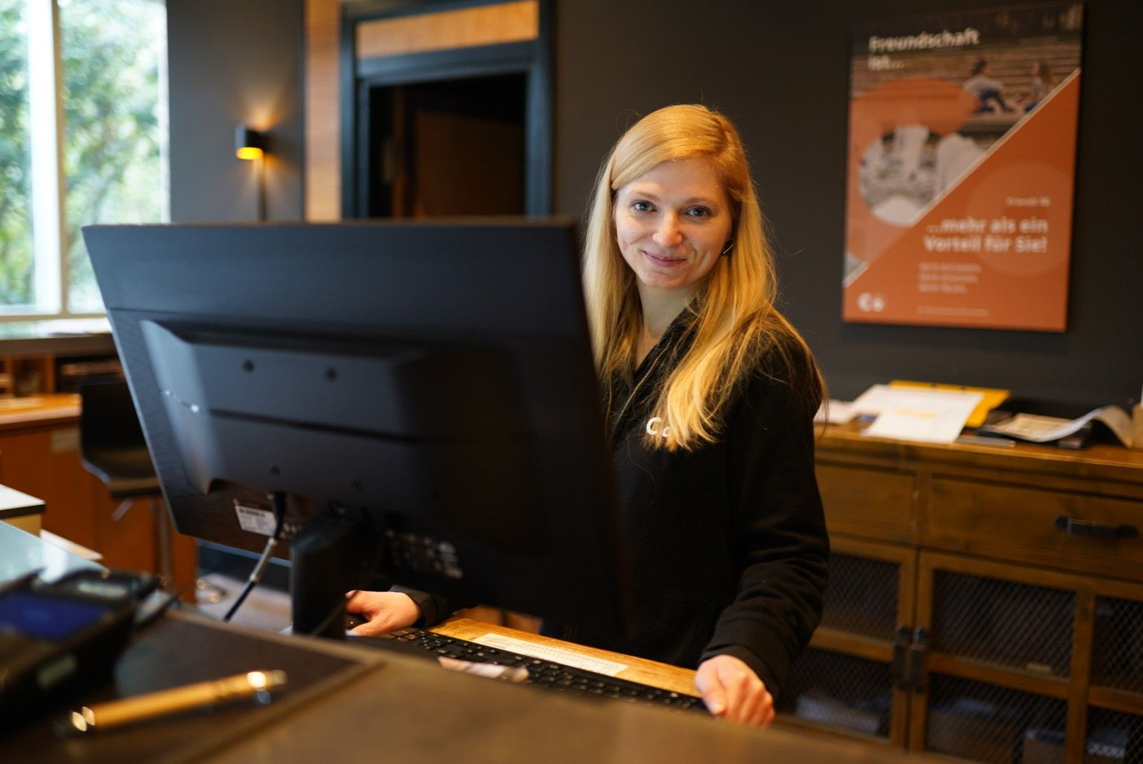 Frau lächelt am Schreibtisch hinter einem großen Monitor und arbeitet. Büroumgebung.