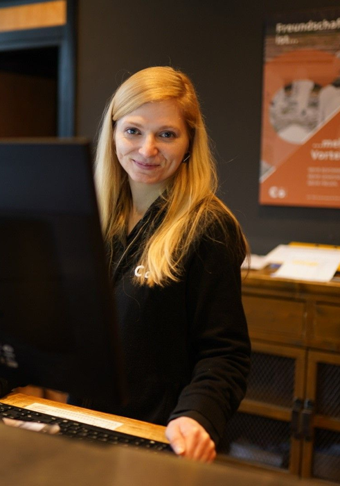 Eine lächelnde Frau steht hinter einem Holztresen in einem schwach beleuchteten Büro oder Empfangsbereich.
