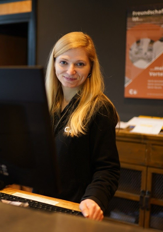 Eine lächelnde Frau steht hinter einem Holztresen in einem schwach beleuchteten Büro oder Empfangsbereich.