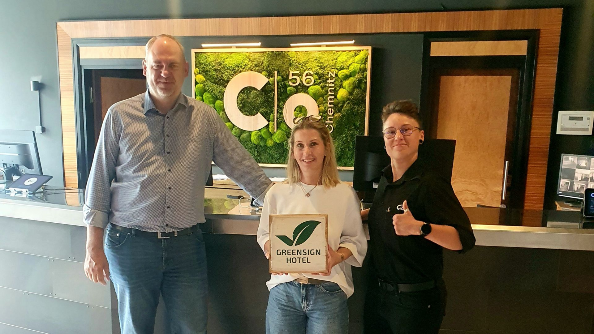 Drei Personen posieren an der Rezeption des c/o56 Chemnitz und haten ein GreenSign Schild.