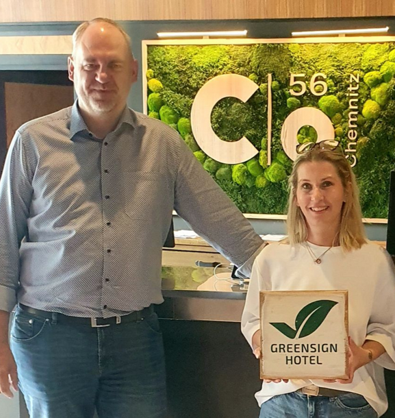 Zwei Personen halten eine GreenSign Hotel-Zertifizierungsplakette vor dem Schild und dem Gebäude des Hotels C 56.