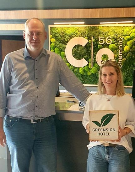 Zwei Personen halten eine GreenSign Hotel-Zertifizierungsplakette vor dem Schild und dem Gebäude des Hotels C 56.