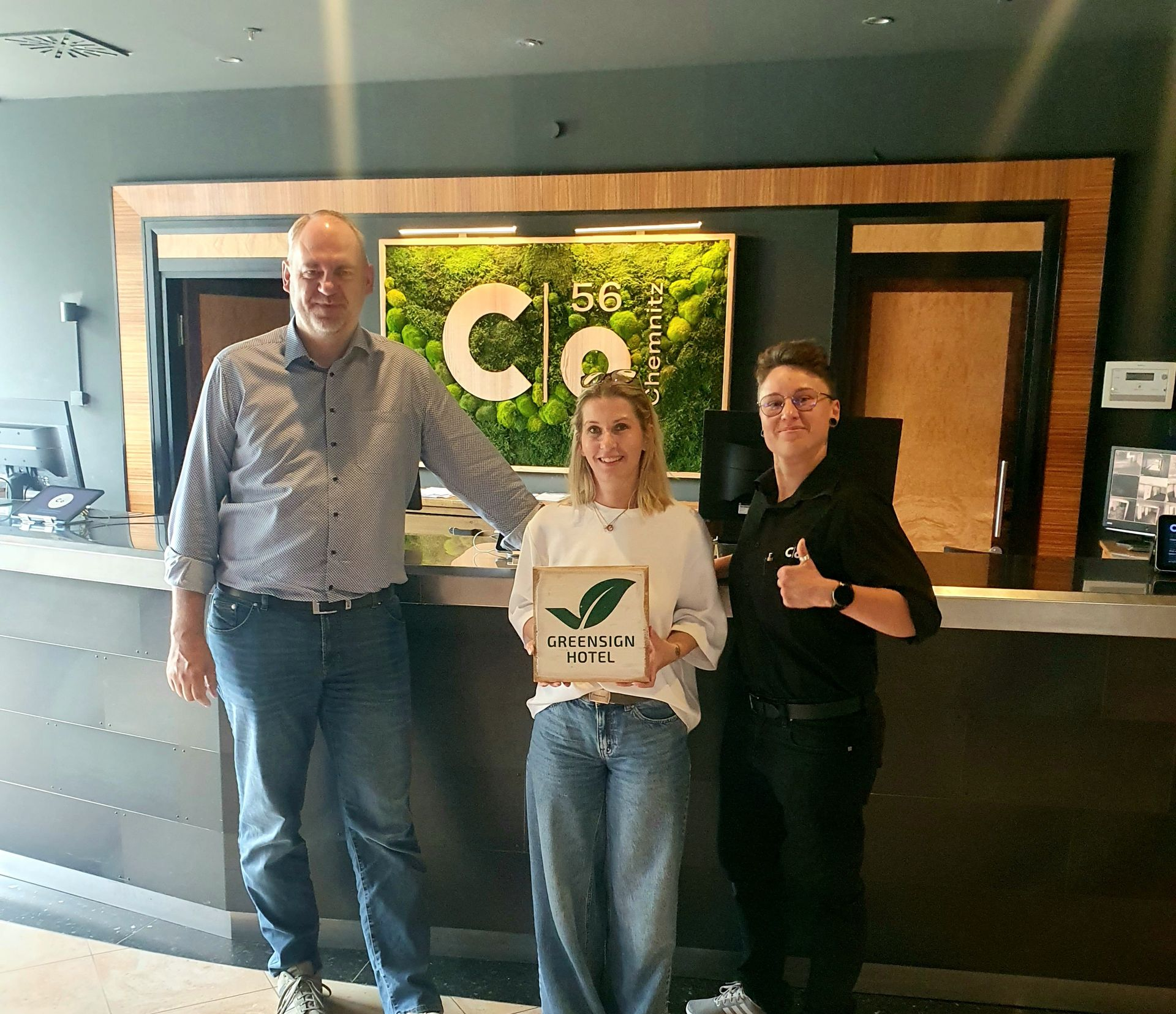 Drei Personen posieren in der c/o56 Chemnitz Lobby und halten die GreenSign Zertifizierung in die Kamera.
