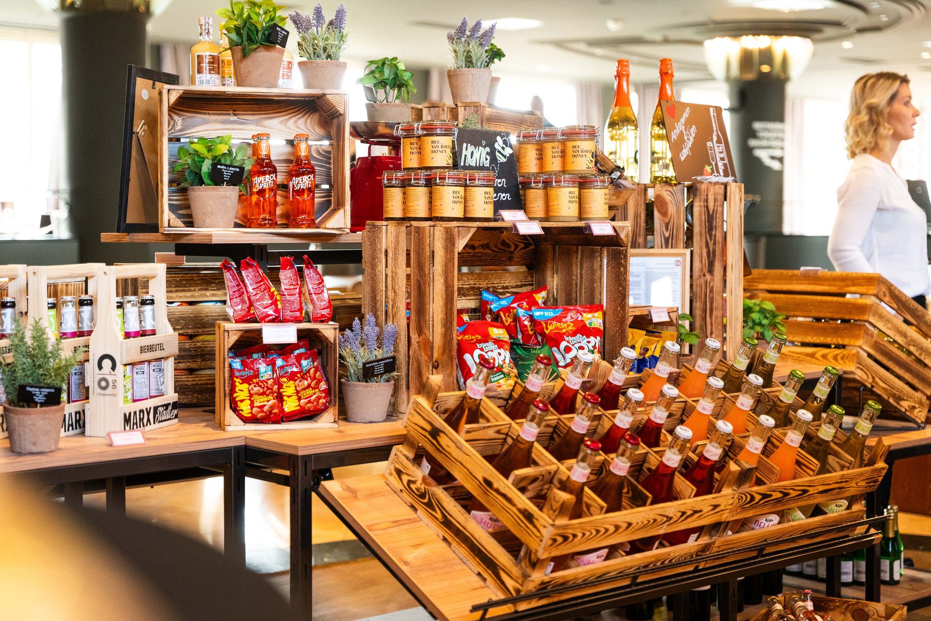 Speisen und Getränke werden in Holzkisten präsentiert. Im Hintergrund steht eine Frau.