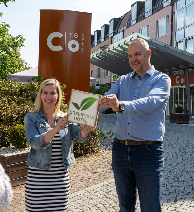 Zwei Personen halten eine GreenSign Hotel-Zertifizierungsplakette vor dem Schild und dem Gebäude des Hotels C 56.