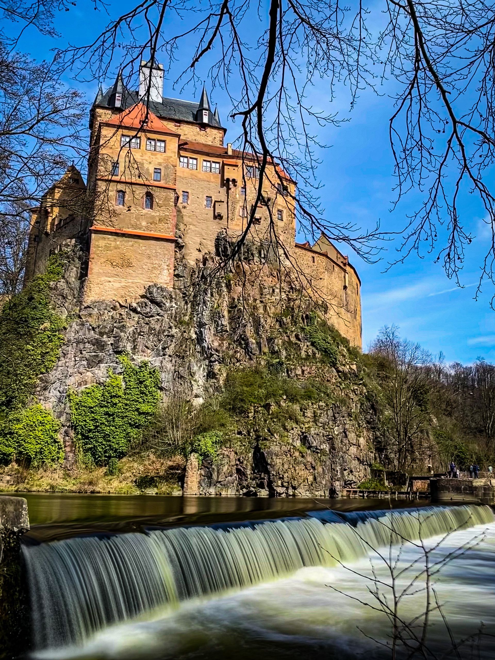 Mittelalterliche Burg, braunes Steingebäude auf einem Felsvorsprung, grüne Bäume, blauer Himmel.