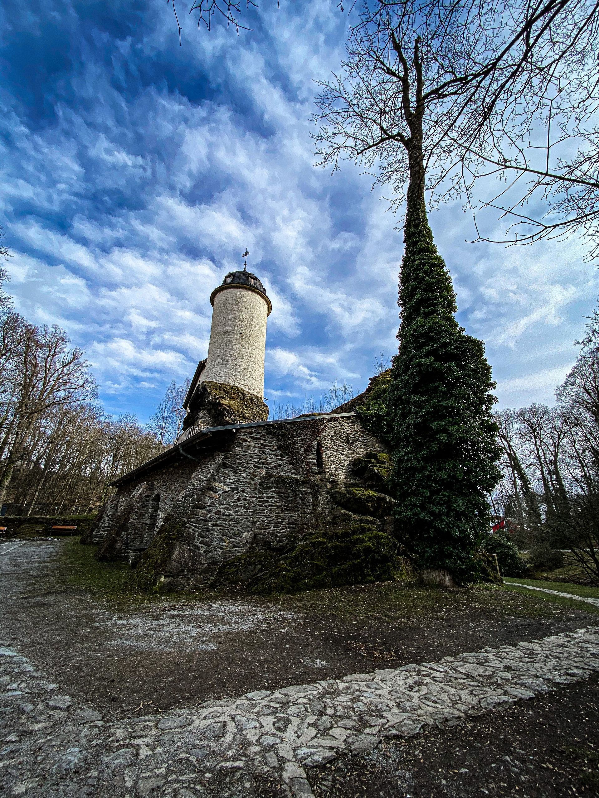 Gelbes Gebäude auf einem grasbewachsenen Hügel mit blauem Zaun. Bäume und blauer Himmel im Hintergrund.