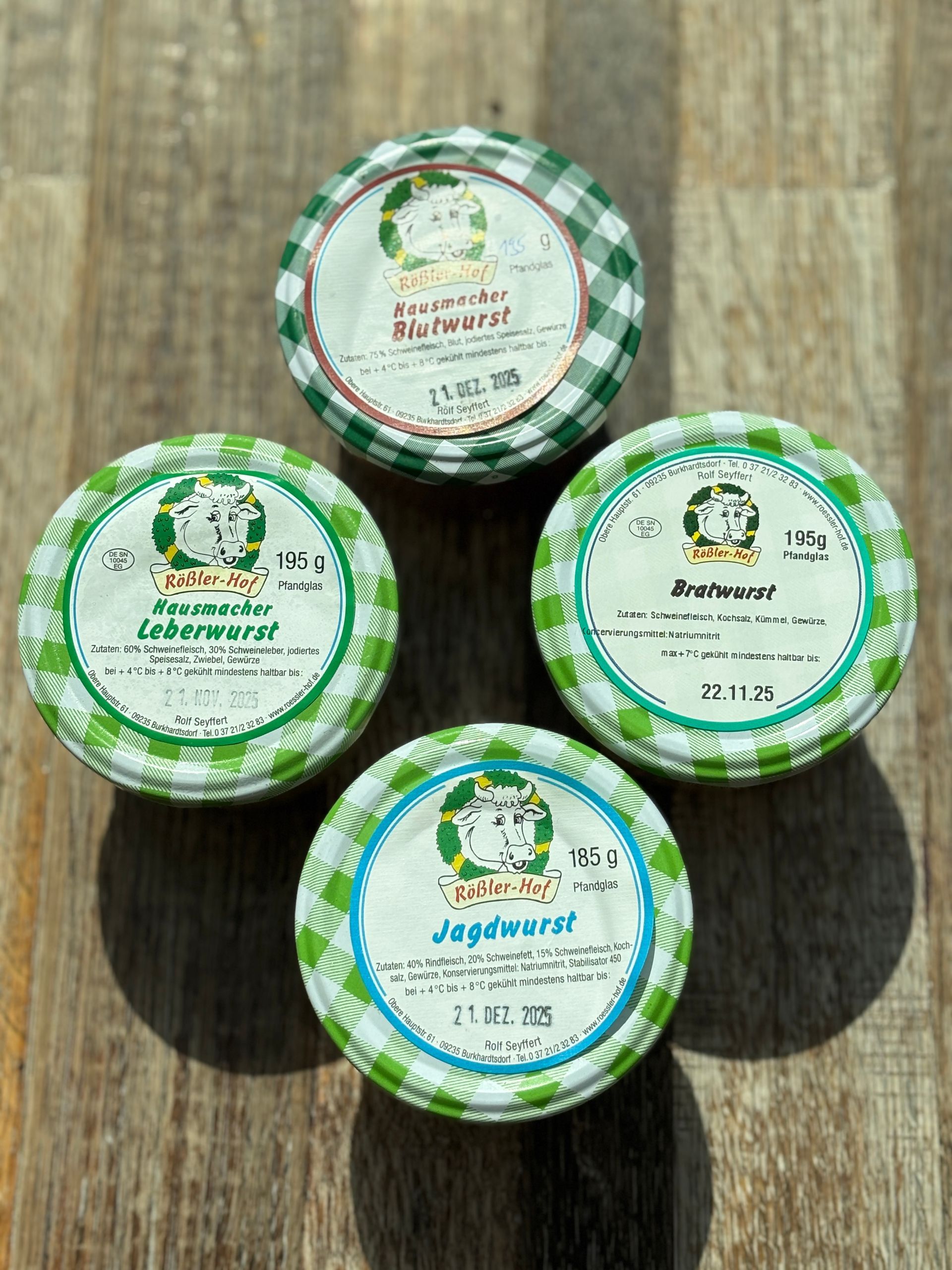 Vier Gläser mit verschiedenen deutschen Wurstsorten und grün karierten Deckeln auf einer Holzoberfläche.