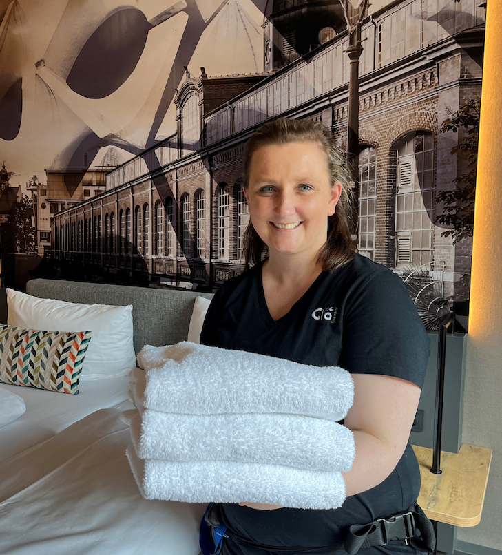 Frau in schwarzer Uniform hält gefaltete weiße Handtücher in einem Hotelzimmer.
