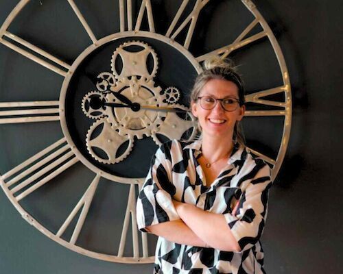 Frau mit Brille lächelt, die Arme verschränkt, steht vor einer großen Uhr mit Zahnrädern.