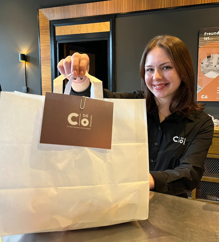 Eine Frau hält in einem Restaurant eine weiße Tüte zum Mitnehmen, an der mit einer Büroklammer ein Firmenlogo befestigt ist.