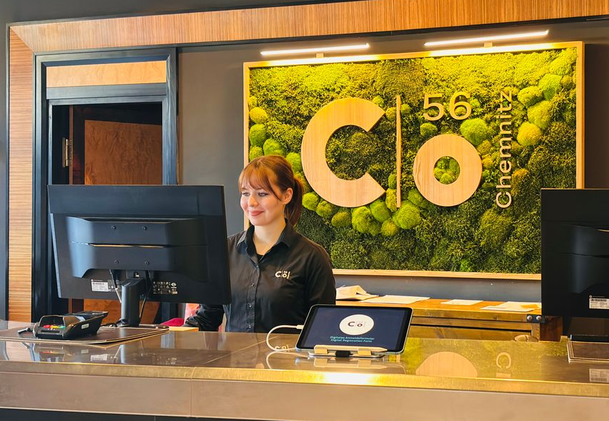 Eine Frau mit Brille und schwarzem Hemd lächelt hinter einem Empfangstresen; das „C“-Logo ist sichtbar.