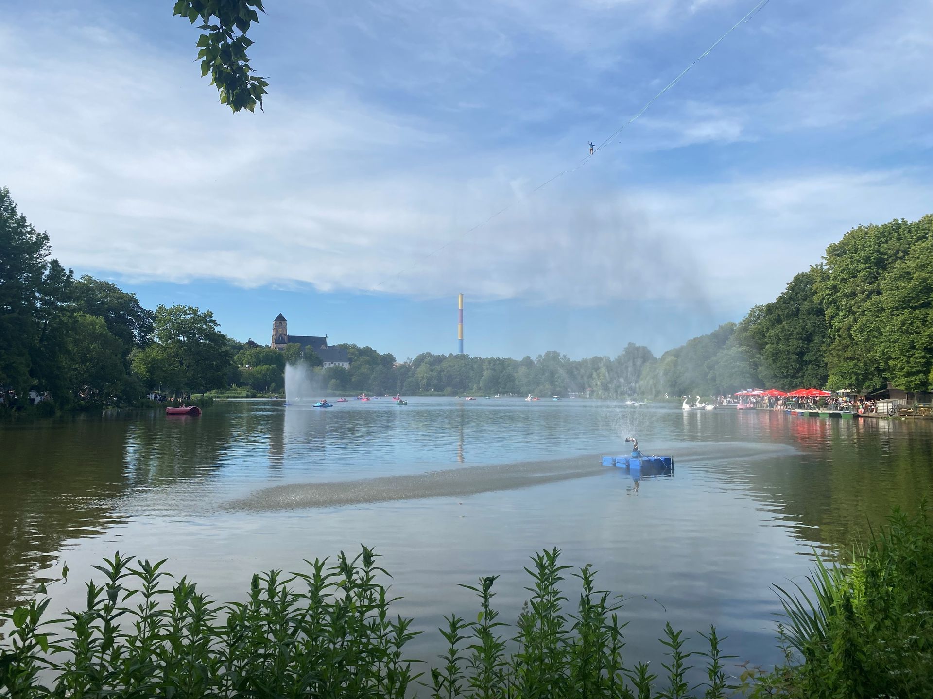 Der Schlossteich mit Springbrunnen und kleinen blauen Boot unter strahlend blauem Himmel.