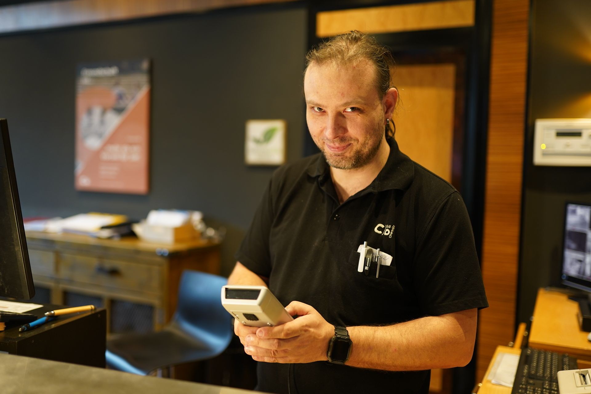 Eine Frau mit Brille und schwarzem Hemd lächelt hinter einem Empfangstresen; das „C“-Logo ist sichtbar.