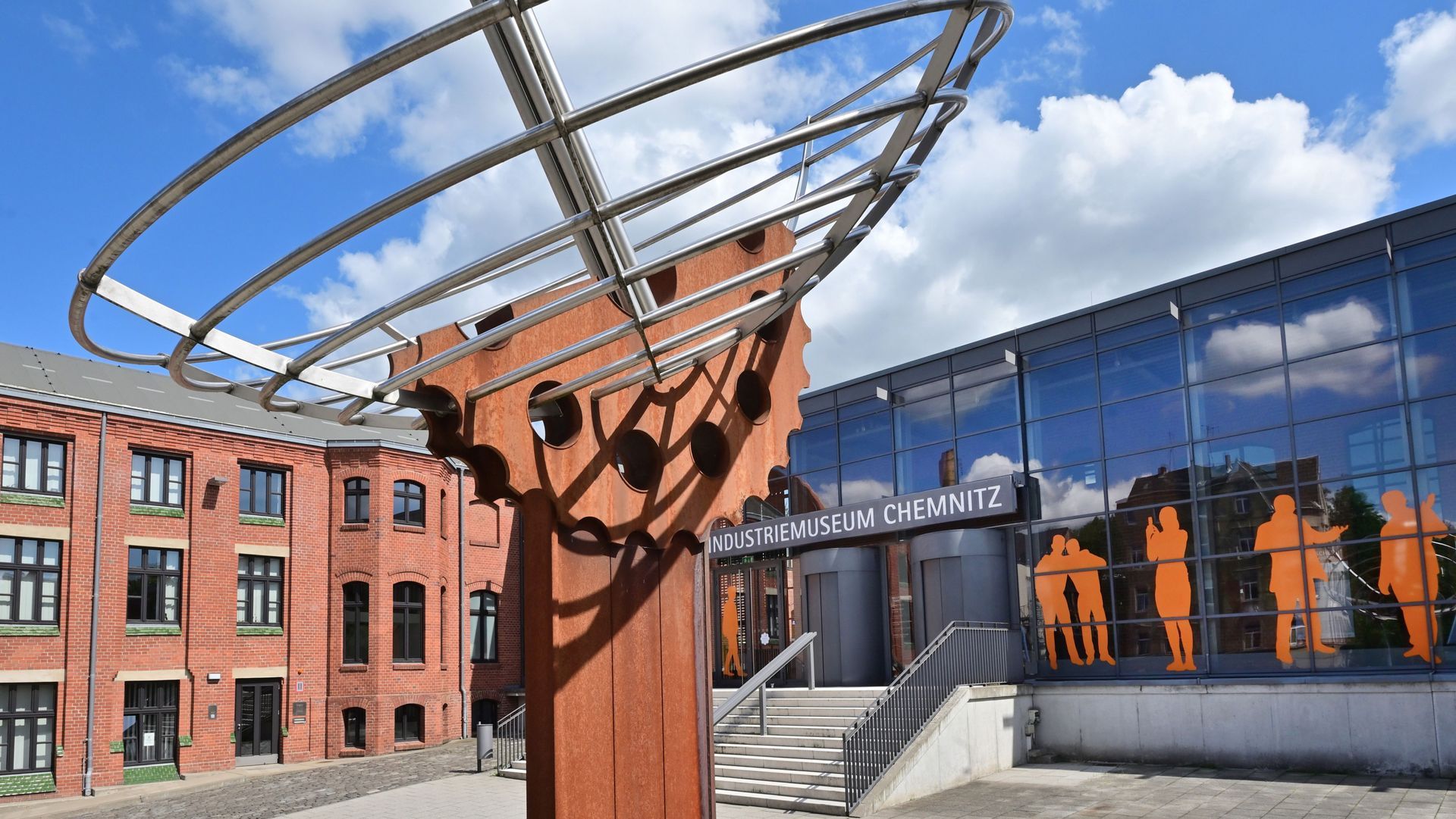 Moderne Skulptur auf einem Platz vor Backstein- und Glasgebäuden, mit orangefarbenen Figuren an einer Wand.