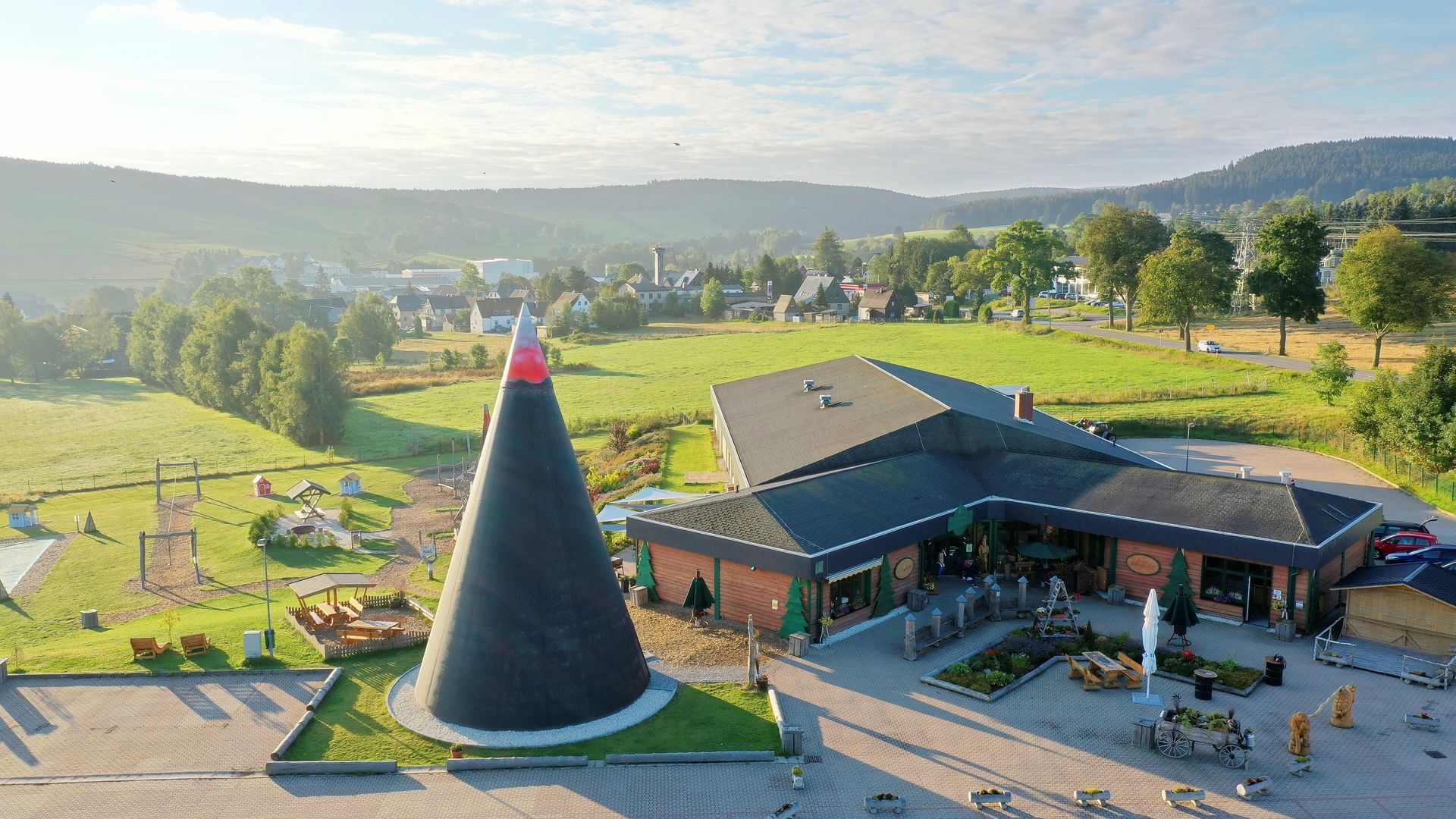 Gelbes Gebäude auf einem grasbewachsenen Hügel mit blauem Zaun. Bäume und blauer Himmel im Hintergrund.